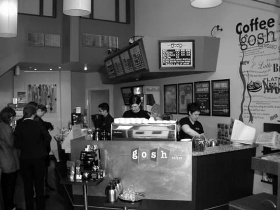 The counter of a GOSH espresso bar run by the founders in the early 2000s, with the coffee menu board and staff making coffee